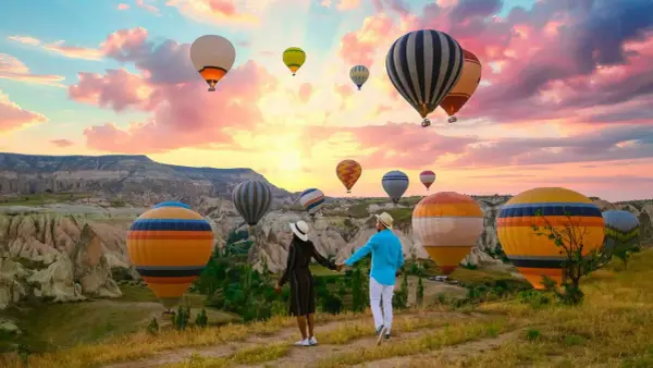 A couple holds hands, gazing at a sky filled with hot air balloons over the unique landscape of western Turkey at sunrise.