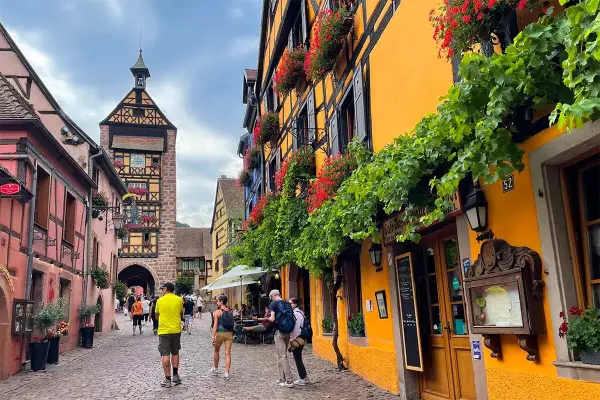 Cobblestone street in a picturesque Alsace town, with colorful half-timbered buildings adorned with flowers. A clock tower stands tall in the background.