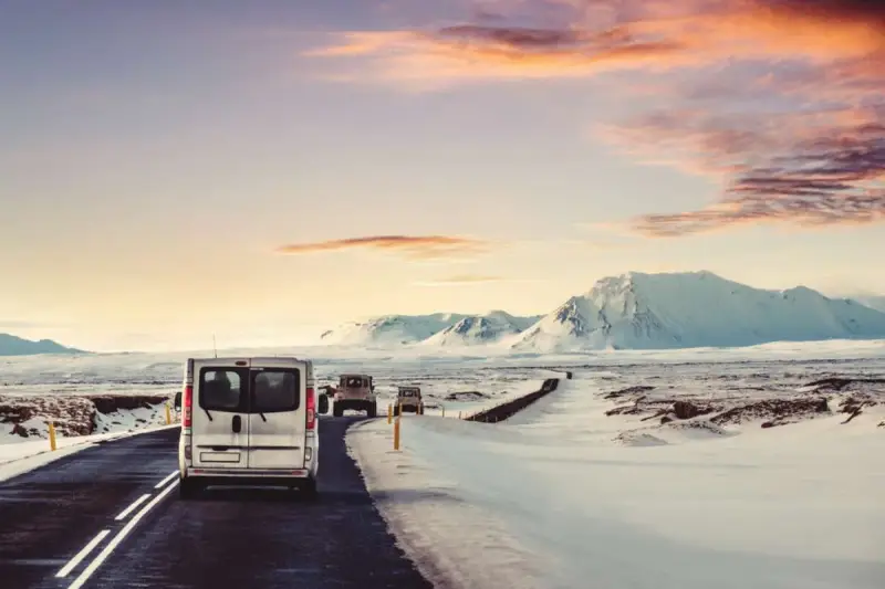 A silver van leads a convoy of vehicles driving in Iceland in December, past snow-covered mountains under a dramatic sunset sky.