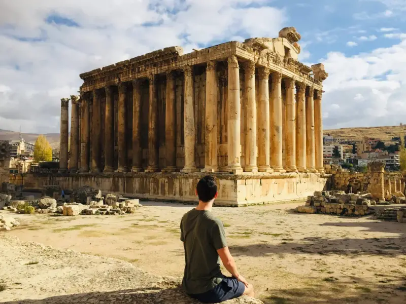 A man sits contemplating the grandeur of Roman ruins in Lebanon, bathed in sunlight under a cloudy sky.