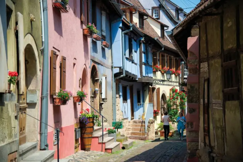 Charming street with colorful houses, perfect for day trips from Strasbourg. Two women stroll past flower-filled window boxes.