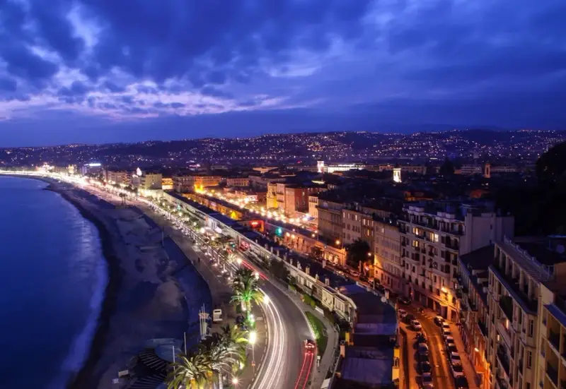 Night view of the Promenade des Anglais from Castle Hill, Nice, France, with city lights twinkling and car trails on the road.
