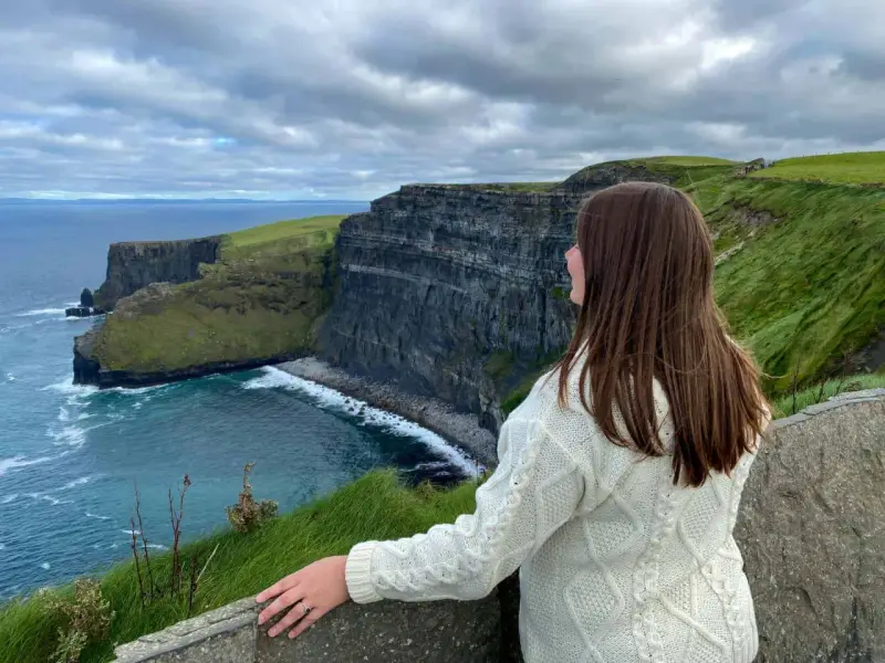 A woman in a cream cable-knit sweater gazes at the dramatic Cliffs of Moher, a breathtaking highlight of many tours of Ireland.