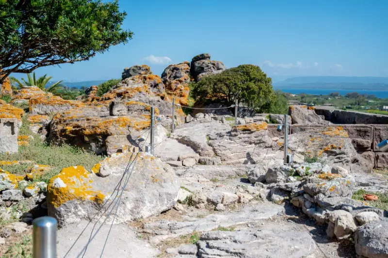 Ancient Italian ruins with weathered stones covered in orange lichen, a pathway winding through the site, and a distant blue sea under a clear sky.